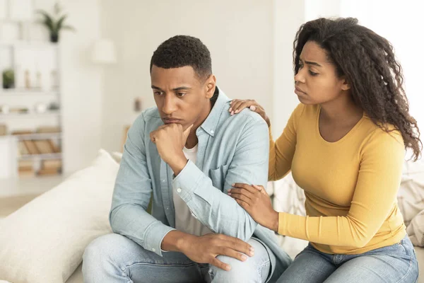 Apologize at home. Sad young african american female calms offended guy, support and caring, sit on sofa in room interior. Sorry after quarrel, relationship problems, bad news reaction and crisis