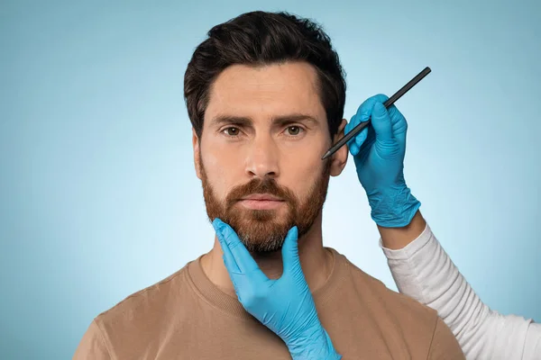 Doctor drawing marks on male face for cosmetic surgery operation over blue studio background. Handsome guy getting ready for aestetic operation or face lifting