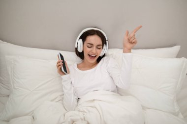 Portrait of happy emotional young brunette lady wearing white pajamas listening to music, singing karaoke and dancing in bed at home, using smartphone and newest wireless headphones
