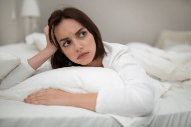 Unhappy young brunette lady in white pajamas laying in bed with open eyes, looking at copy space, having bad days, bedroom interior, closeup shot. Depression, frustration, loneliness concept