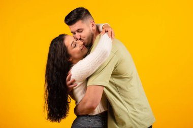 Satisfied handsome millennial middle eastern man hugging and kissing female, couple enjoy free time and tender moment date isolated on orange background, studio. Family relationships, love and romance
