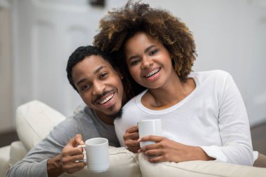 Beautiful cheerful happy loving black spouses drinking coffe at home, smiling african american young man and woman sit on couch in living room, cuddling and enjoying tea, family portrait, copy space