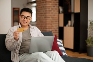 E-commerce concept. Happy asian mature man holding credit card and shopping online with laptop computer, sitting on sofa at home, free space. Male customer showing bankcard