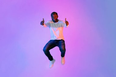 Cool happy handsome mature african american man in casual outfit jumping up and showing thumb ups on colorful neon studio background, copy space for advertisement, full length