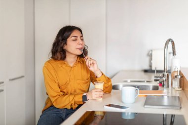 Young Arab Woman Sitting At Kitchen Counter And Eating Tasty Pudding From Plastic Cup, Happy Millennial Middle Eastern Female Licking Spoon While Enjoying Delicious Dessert Meal At Home, Copy Space