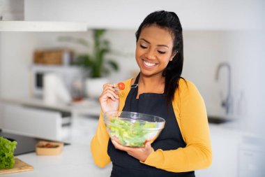 Smiling young african american female chef in apron eating organic vegetable salad, enjoy fresh dish in modern kitchen interior. Delicious diet, lose weight and cooking healthy homemade food at home