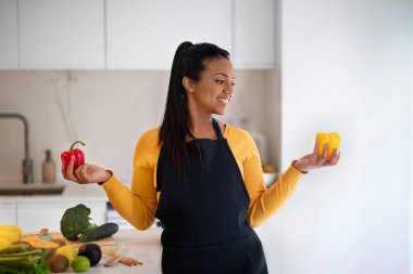Happy young african american woman in apron hold red and yellow pepper in hands at table with organic vegetables in modern kitchen interior. Food choice, cooking homemade eat at home, health care