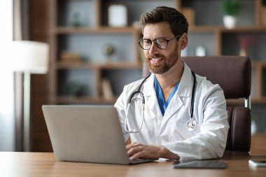 Smiling Handsome Male Doctor Working On Laptop While Sitting At Desk In Office, Happy Physician Man Wearing Medical Coat Consulting Patients Online, Typing On Computer Keyboard, Copy Space