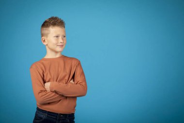 Portrait Of Handsome Preteen Boy With Folded Arms Standing Over Blue Background, Pensive Male Child Looking Away And Smiling, Thinking About Something While Posing In Studio, Copy Space