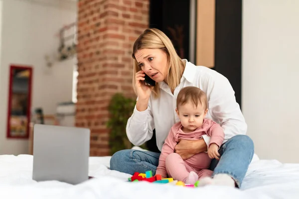 Busy mom working distantly and caring for child during maternity leave, woman using laptop and talking on phone, sitting with baby toddler on bed at home, free space. Freelance job and motherhood