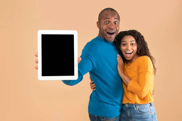 Happy black spouses showing digital tablet with black screen, family recommending new app or cool website, posing on peach studio background, mockup