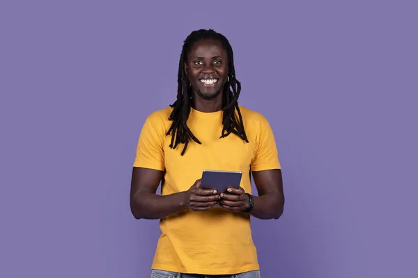 在紫色工作室背景上使用现代数码垫 查看最新的娱乐性移动应用程序 复制空间 快乐地笑着 留着长发 穿着休闲装的年轻黑人小伙子 — 图库照片
