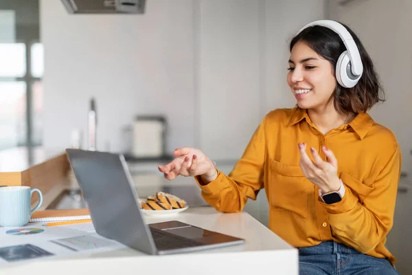 Video Call. Happy Millennial Arab Woman In Headphones Teleconferencing On Laptop In Kitchen, Smiling Middle Eastern Female Freelancer Talking And Gesturing At Computer Web Camera, Free Space