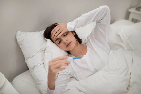 Young sick brunette woman with fever checking her temperature with thermometer and touching her forehead in bed at home, lady got sick, copy space. Cold, flu, coronavirus concept
