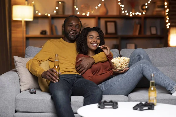Cheerful multi generational black spouses chilling together at home during Christmas holidays, happy man and woman sitting on couch in living room decorated with lights, watching movie, drinking beer