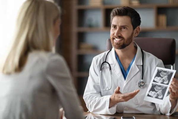 Gynecologist Doctor Consulting Pregnant Female Patient In Clinic, Showing Baby Sonography Image To Young Woman During Meeting In Office, Smiling Reproductive Endocrinologist Consulting Lady