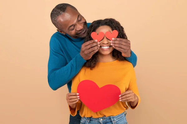 情人节庆祝的概念 可爱的中年黑人男人用纸心盖住妻子的眼睛 摆出桃子工作室的样子 这是我的情人节观念 — 图库照片