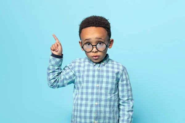 Look here. Little black boy in eyeglasses pointing aside, showing free copy space for ad, looking at camera over blue background, panorama, studio shot