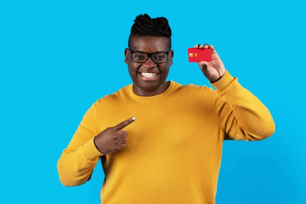 Cheerful Black Man Pointing At Credit Card In His Hand And Looking At Camera, Happy Young African American Male Recommending Bank Services While Standing Over Blue Studio Background, Copy Space