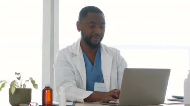 Young professional african american man doctor in robe working on laptop at office, smiling to camera at workplace, tracking shot, slow motion, empty space