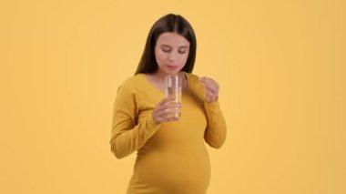 Healthcare during pregnancy. Young pretty pregnant woman taking vitamin pill and drinking water, smiling to camera, orange studio background, slow motion, free space