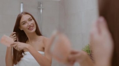 Morning beauty habits. Mirror reflection portrait of young positive woman wrapped in towel brushing her hair and bathroom at home, tracking shot, slow motion, empty space