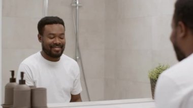 Good morning routine. Young positive african american guy looking at mirror and smiling to himself at bathroom after washing in bathroom, tracking shot, slow motion, free space