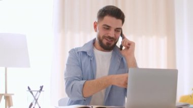 Telemarketing concept. Young positive arab guy sales agent consulting client via cellphone and looking at laptop for information, working at home office, tracking shot, slow motion, free space