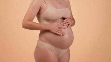 Skin care during pregnancy. Unrecognizable pregnant woman in underwear rubbing pampering cream heart on her belly, beige studio background, slow motion