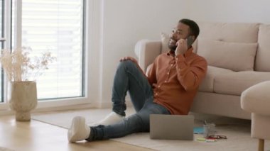 Distant business negotiations. Young carefree african american male manager talking to partners on phone, working on laptop from home, sitting on floor, tracking shot, slow motion, free space