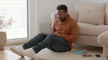 Comfort of freelance. Young positive african american man typing project on laptop, sitting on floor at home interior, tracking shot, slow motion, empty space