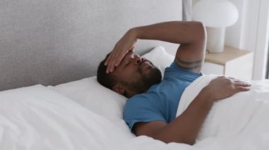 Bad sleep problem. Young african american guy waking up in bed and massaging forehead, suffering from headache and keep resting, tracking shot, slow motion, empty space