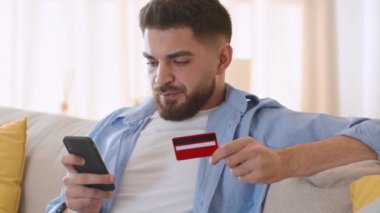 Mobile banking app. Young concentrated arab guy making online financial payment with credit card and smartphone, sitting at home, tracking shot, slow motion, free space