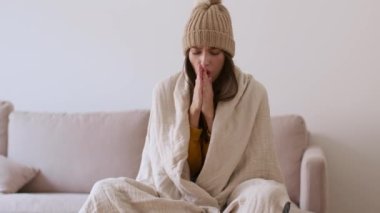 Apartment heating problem. Young unhappy woman freezing at home, sitting in knitted warm hat and wrapping herself in plaid, shivering on sofa, tracking shot, slow motion, free space