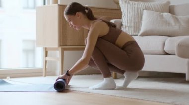 Done training. Young sporty woman rolling fitness mat at home after finishing fitness workout, side view, slow motion, empty space