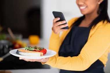 Millennial african american woman in apron taking photo of fish on phone for blog and social media in modern kitchen interior, close up. Work of professional chef, cook healthy eat at home, food blog