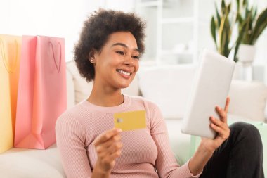 Smiling young mixed race female shopaholic with tablet, credit card checking financial account, use banking app in room interior with many bags from store. Sale and cashback, online shopping at home