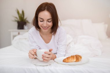 Candid photo of cheerful happy beautiful brunette young woman in white pajamas have breakfast and smiling, lady drinking coffee, eating croissant in bed after waking up in the morning, copy space