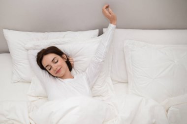 Happy pretty brunette young woman wearing white long sleeve pajams stretching in bed with closed eyes and smile on her face after waking up in the morning at home, copy space next to female