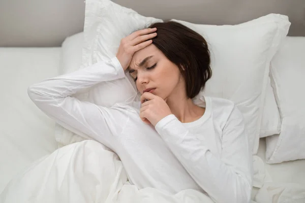 Top view of unhappy young brunette woman lying in bed at home, touching head and chin, tired lady wake up with headache in the morning, suffering from chronic migraine, copy space