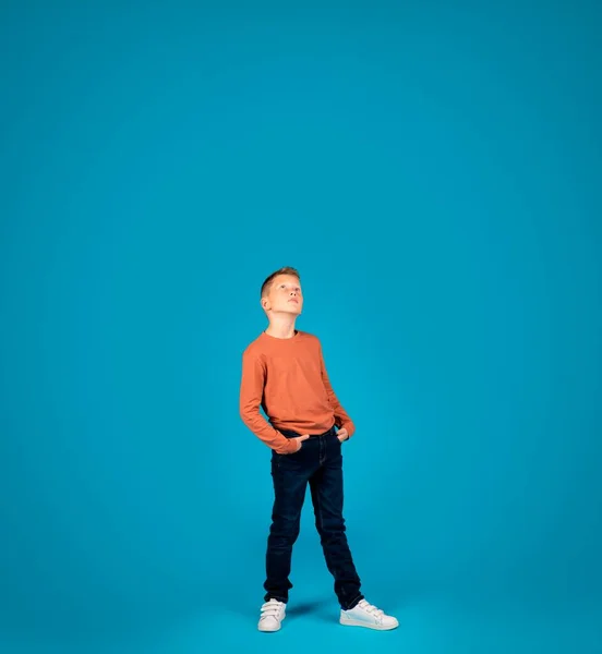 Curious Preteen Boy Looking Up With Interest While Standing Over Blue Background In Studio, Dreamy Male Child Thinking About Something, Glancing At Copy Space Above His Head, Full Length Shot