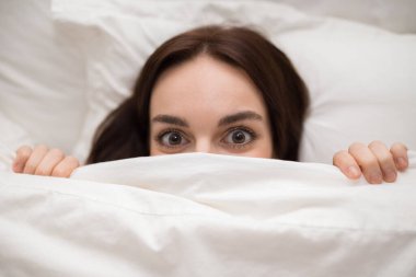 Closeup shot above of young brunette woman hiding under duvet blanket in white bed, showing beautiful big brown eyes, playful lady having fun while chilling in bedroom at home