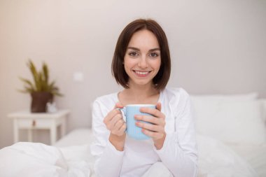 Positive attractive brunette young lady wearing white cotton pajamas sitting alone in bed, holding blue mug and smiling at camera, female enjoying fresh morning coffee at home, copy space