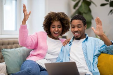 Lottery, giveaway, betting. Emotional happy beautiful african american young couple gambling on Internet at home, cheerful black lovers winners sitting on sofa, using laptop and rasising hands up