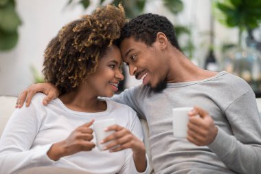 Portrait of sweet cheerful loving african american couple sitting on sofa in living room at home, embracing, cuddling, touching with foreheads, chatting while drinking coffee, closeup