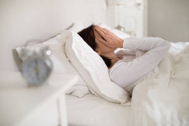 Tired young brunette woman in white pajamas lying in bed at home in the morning, touching her face with both hands, wake up early, blurred mechanical alarm on bedside table, side view