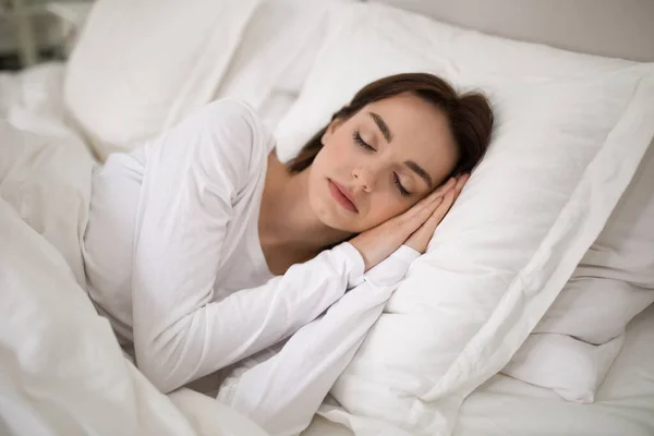 Young beautiful brunette lady sleeping with hands under her head on soft comfy pillows, peaceful woman lying in bed with closed eyes, candid photo. Resting, sleeping, relaxation at home concept