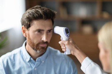 Doctor Lady With Infrared Thermometer Checking Body Temperature For Male Patient In Clinic, Sick Middle Aged Man Getting Checkup In Hospital During Covid-19 Pandemic, Selective Focus