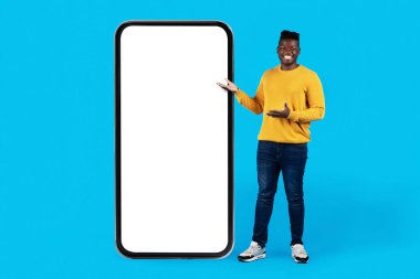 Check This. Cheerful Black Man Standing And Pointing At Big Blank Smartphone With White Screen, Happy African American Guy Advertising New Mobile App Or Website, Standing Over Blue Background, Mockup