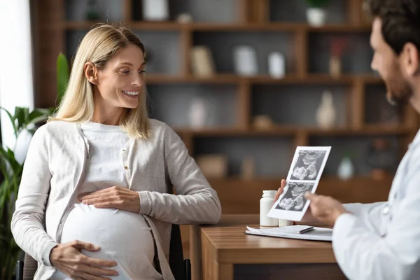 院内集会中の若い妊婦にベビーソノグラフィー画像を見せる婦人科医 笑顔で生殖器科医コンサルティング病院での幸せな妊婦 フリースペース — ストック写真